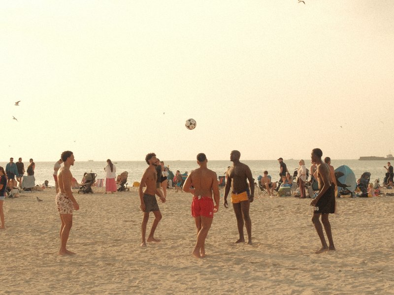 Group of people playing beach volleyball on a sandy beach with a hazy sky and distant beachgoers.