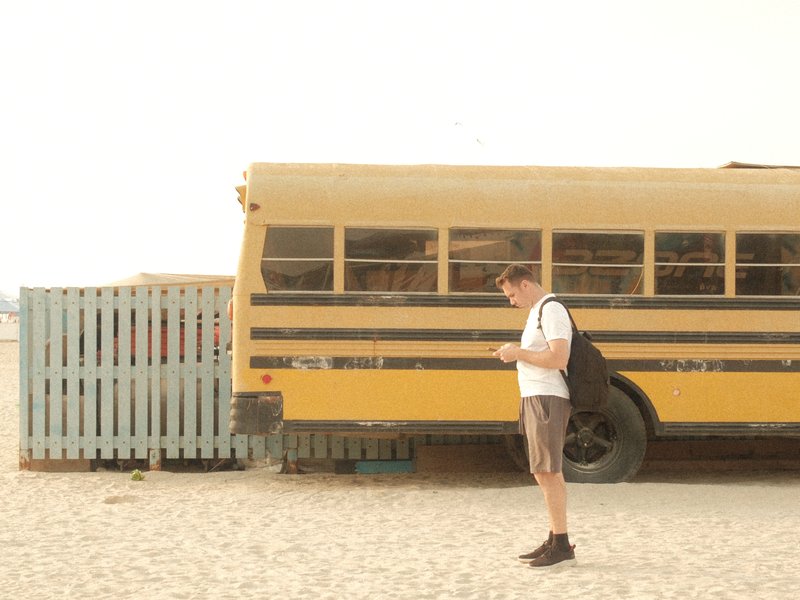 A young man standing beside a yellow school bus on a sandy beach, checking his phone.