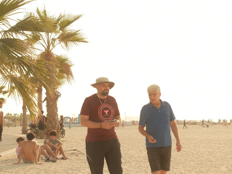 Two men walking on a beach with palm trees, soft sunlight creating a warm atmosphere.