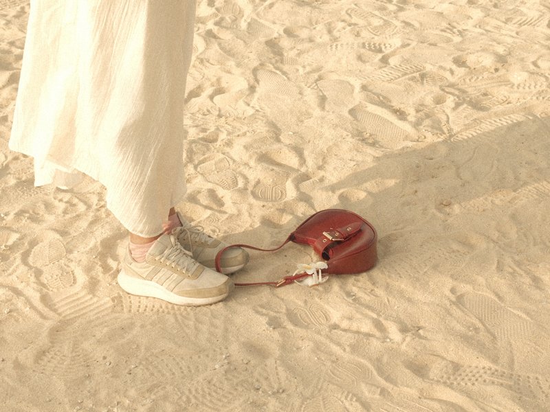 A person's foot in sneakers next to a small red handbag on sandy ground.
