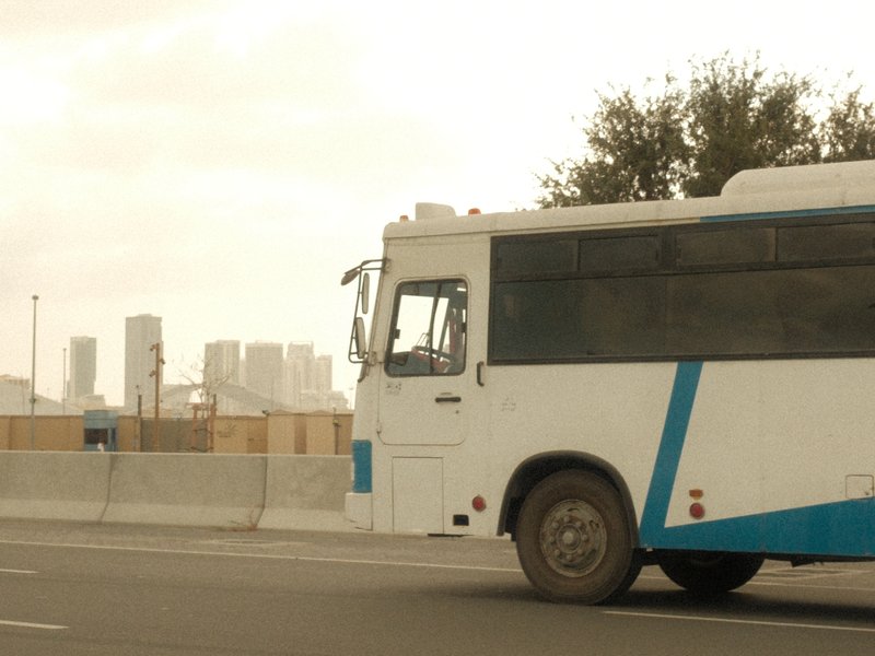 A white and blue bus parked on a road with a city skyline in the background under a cloudy sky.