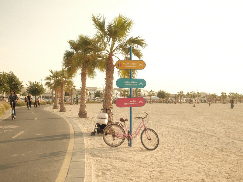 A pink bicycle parked beside a directional sign on a sandy beach with palm trees and people in the background.