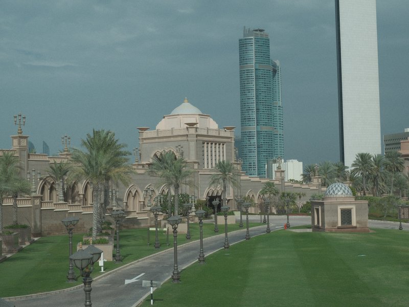 A view of a grand building with palm trees and modern skyscrapers in the background under a cloudy sky.