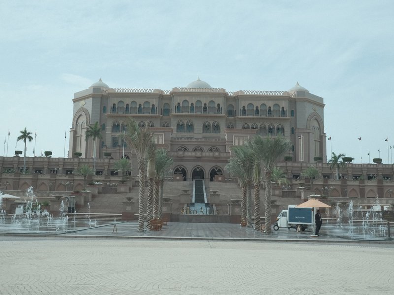 A grand palace with a large staircase and palm trees in front, under a cloudy sky.