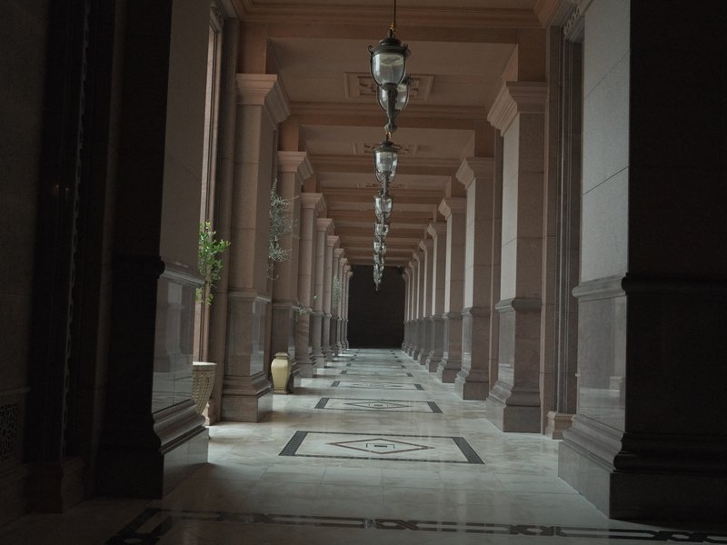 A long corridor with columns and lanterns, leading to a dark end, featuring marble flooring and a plant.