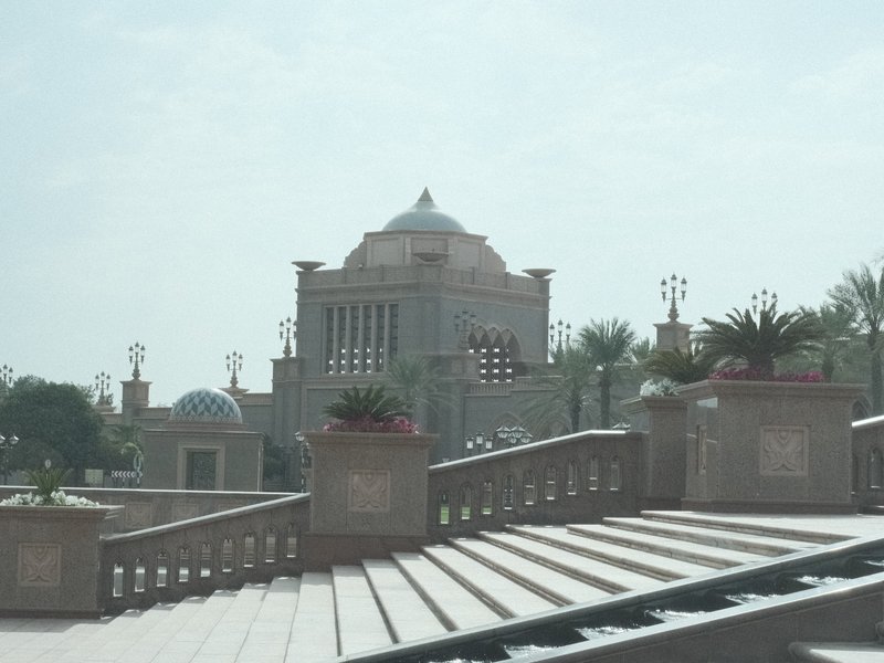 A grand building with a dome and decorative elements, surrounded by palm trees and a staircase.
