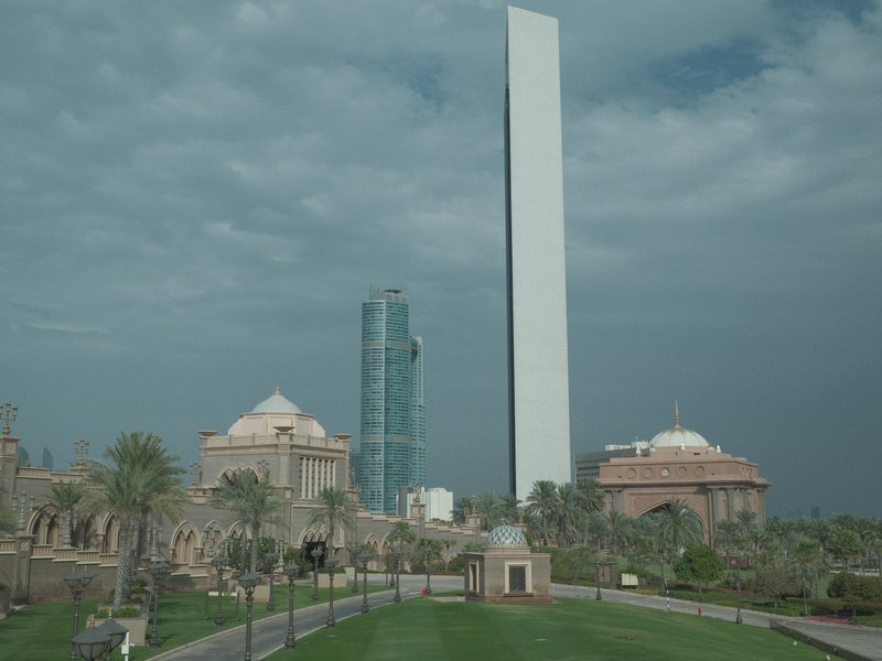 A modern skyscraper towers over traditional architecture in a park setting under a cloudy sky.