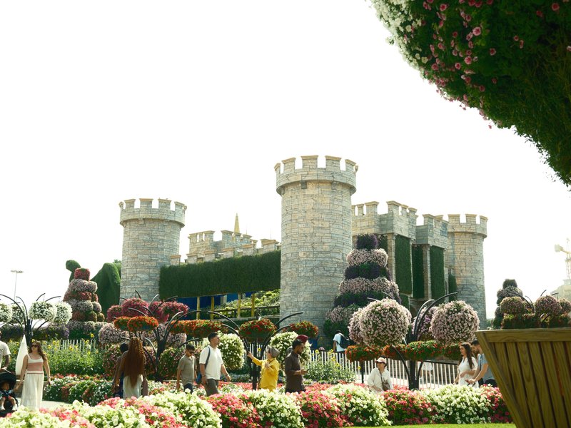 A stone castle surrounded by colorful flower arrangements and visitors in a bright outdoor setting.