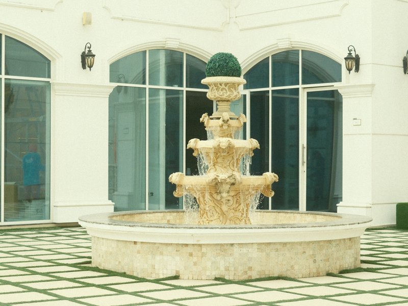 A decorative stone fountain with multiple tiers, surrounded by a tiled courtyard and large glass windows.