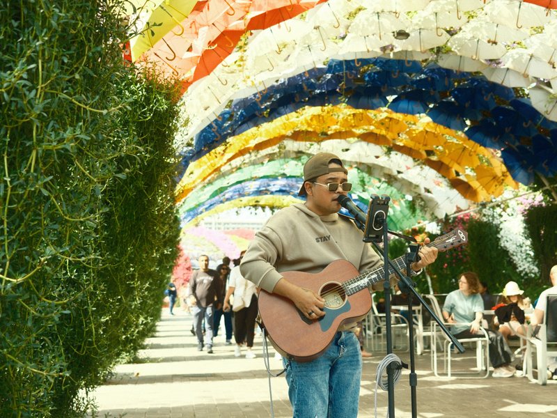 A musician playing guitar under a colorful archway with people in the background.