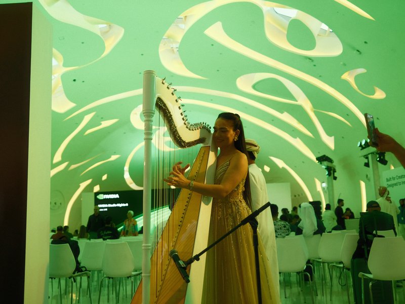 A woman playing a harp under a green-lit ceiling with abstract patterns in a modern indoor setting.