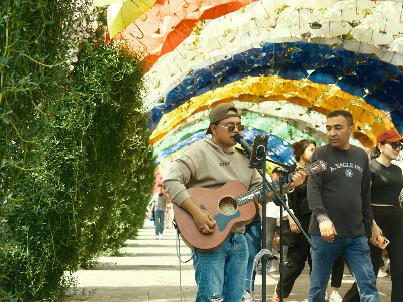 A musician playing guitar under a colorful archway with people walking by.