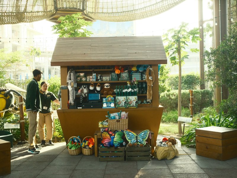A small wooden kiosk filled with colorful items, surrounded by greenery and two people interacting.