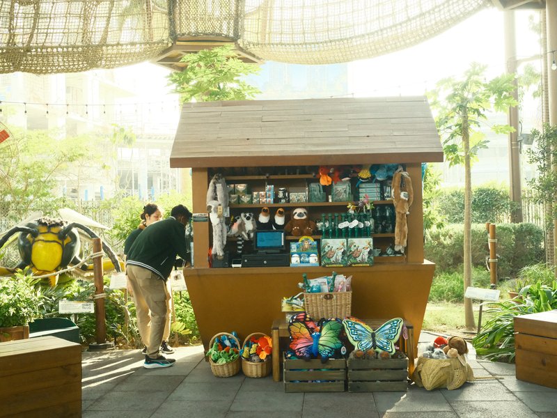 A small wooden kiosk filled with colorful items, surrounded by greenery and sunlight.