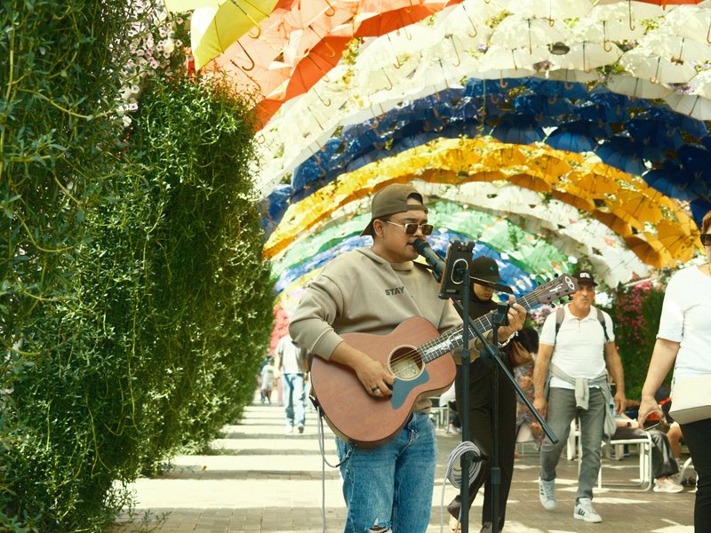 A street musician playing guitar under a colorful archway with people walking by.