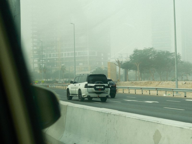 A white SUV driving on a foggy highway with blurred buildings in the background.