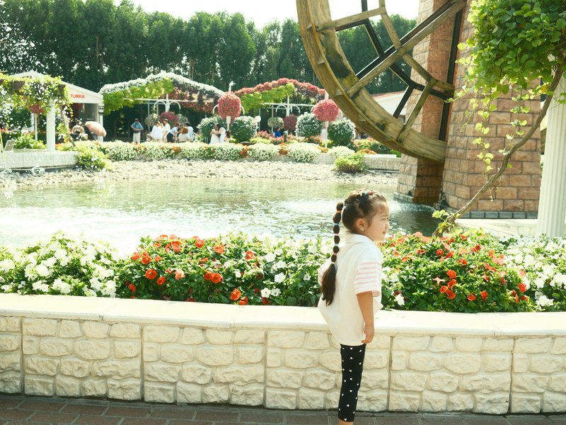A young girl stands by a flower bed in a garden with a water wheel and people in the background.