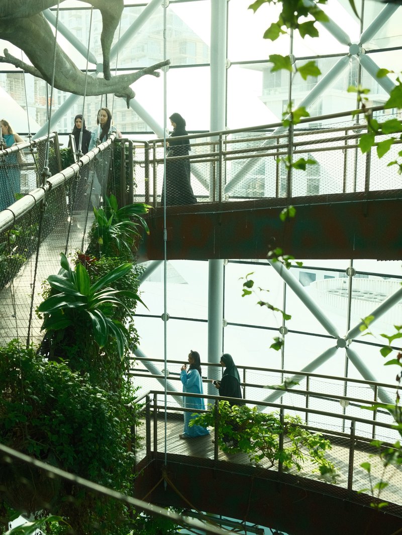 People walking on a wooden bridge surrounded by greenery in a modern glass structure.