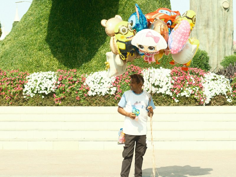 A balloon vendor stands in front of colorful flower beds, holding a bunch of character-themed balloons.