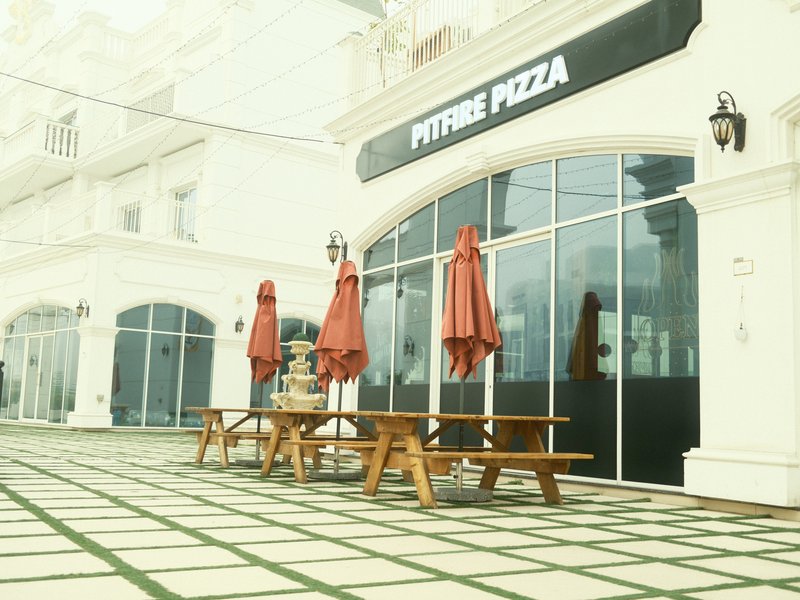 Outdoor seating area of a pizza restaurant with red umbrellas and a tiled floor.