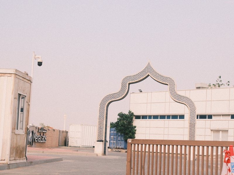 A modern archway leading to a building with a white facade, surrounded by a dusty environment.