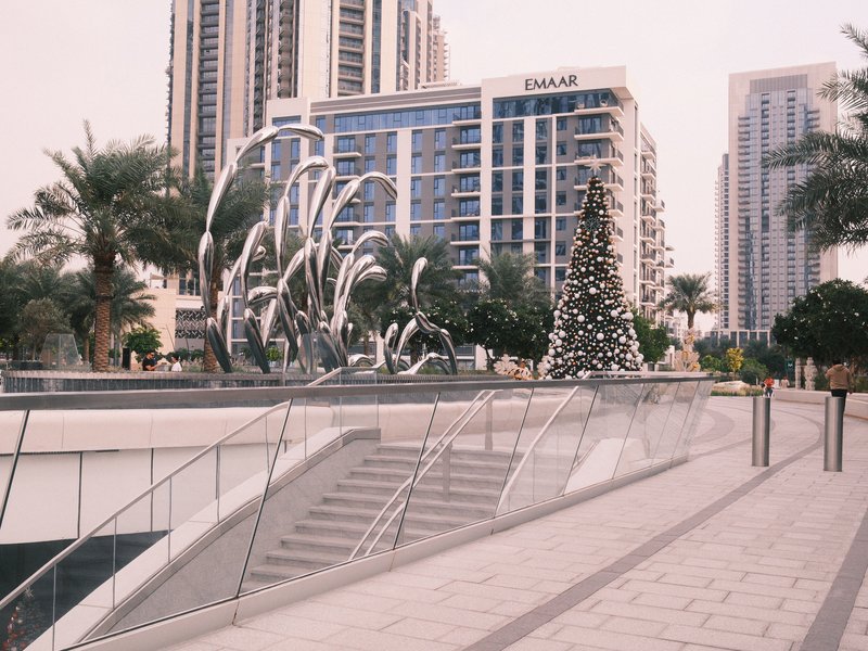 A modern urban scene featuring a decorative fountain and a Christmas tree amidst tall buildings.