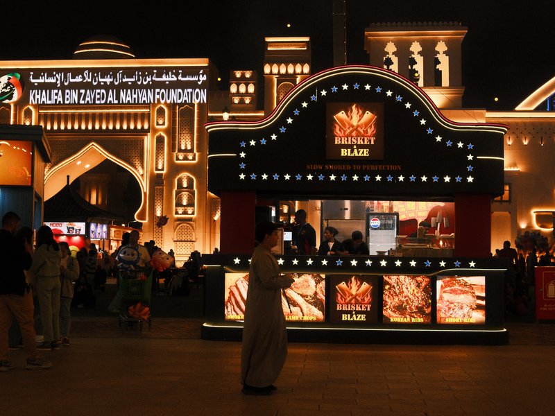 A vibrant food stall illuminated at night with decorative lights and a crowd in the background.