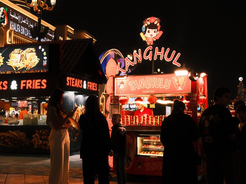 A vibrant food stall at night with people gathered around, illuminated by colorful lights.