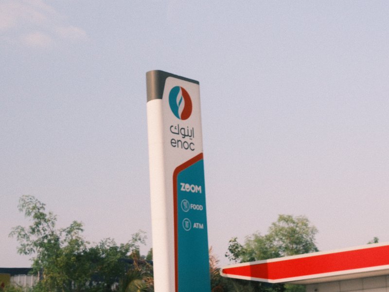 A gas station sign with a clear sky in the background, featuring green trees and a red roof.