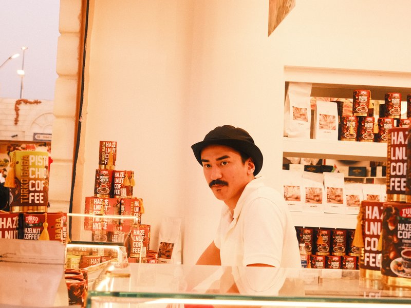 A man in a hat stands behind a counter filled with coffee cans in a bright shop.