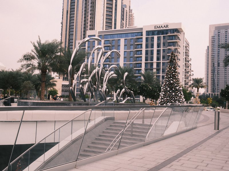 A modern urban scene featuring metallic sculptures and a decorated Christmas tree amidst tall buildings.