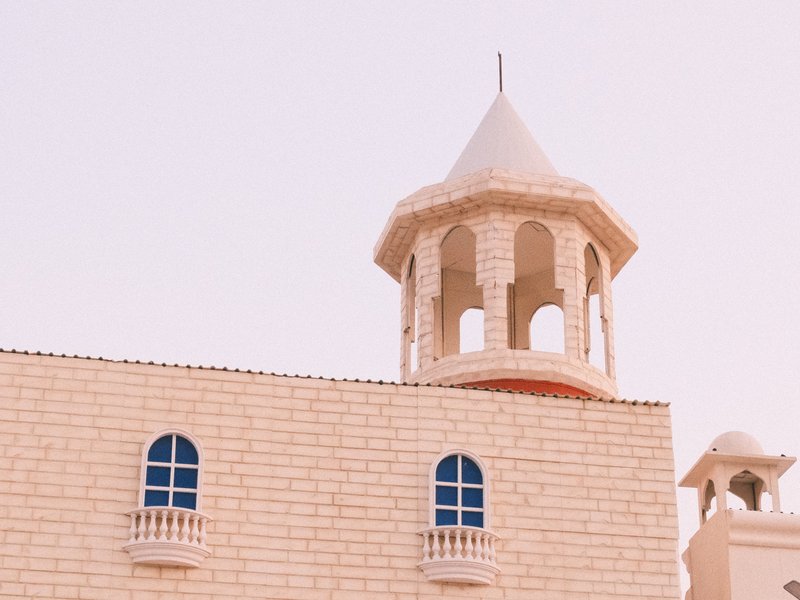 Close-up of a stone building with a tower and arched windows against a pastel sky.