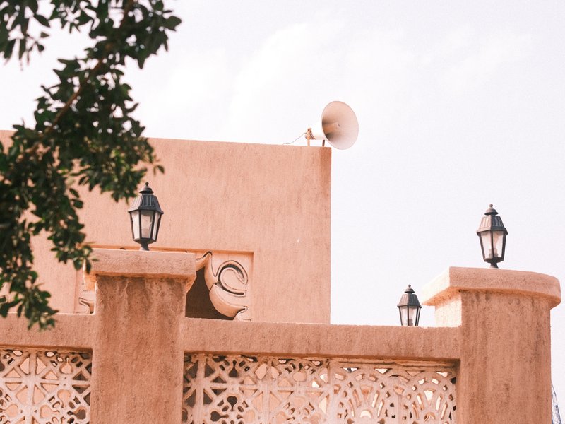 A close-up view of a textured wall with decorative patterns, lanterns, and a satellite dish against a pale sky.