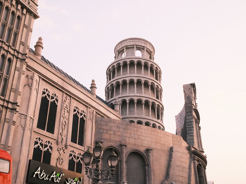 A replica of the Leaning Tower of Pisa next to a detailed architectural building under a pastel sky.