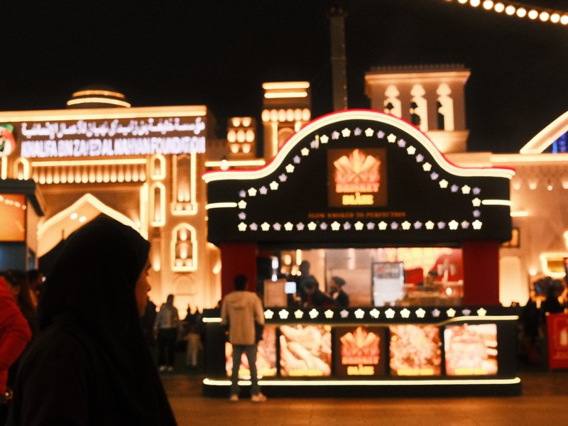A vibrant night scene at a festival with illuminated food stalls and a person in traditional attire.