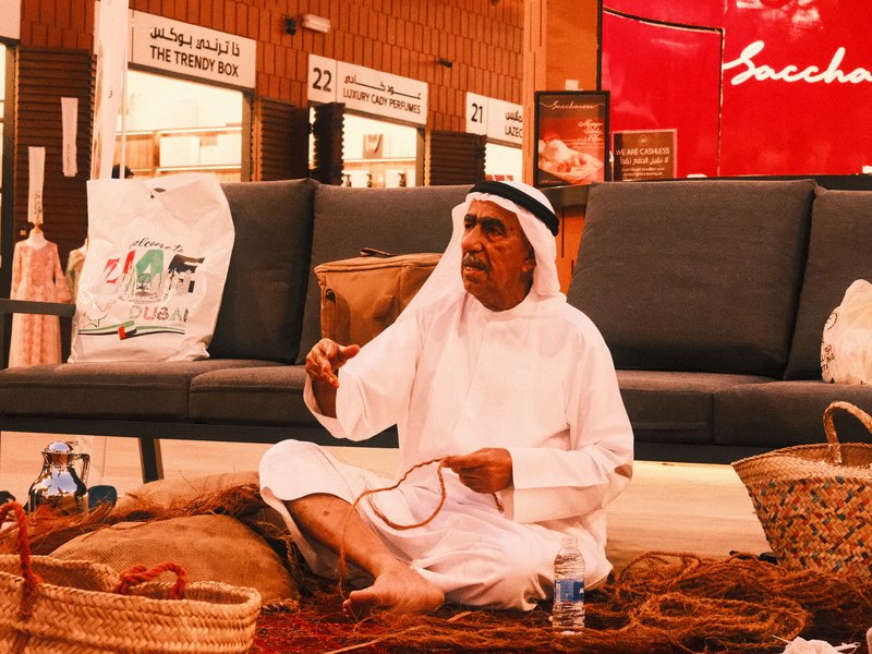A man in traditional attire sitting on the floor of a market, engaged in conversation with a woven basket nearby.
