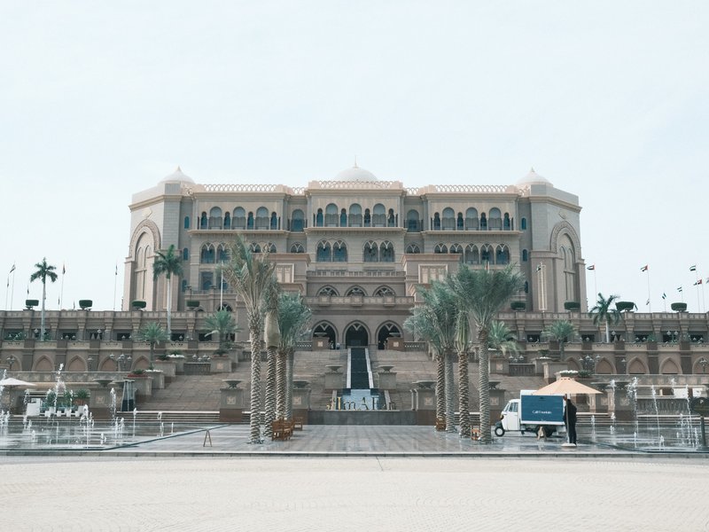 A grand palace with a large staircase and palm trees in front, under a bright sky.
