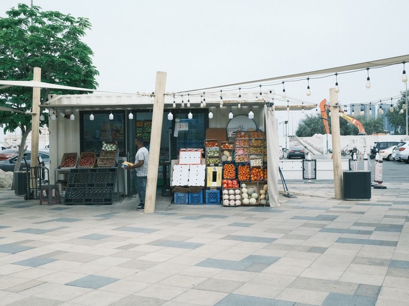 A fruit stall with crates of produce in a market setting, surrounded by a modern urban environment.