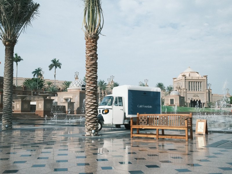 A white delivery truck parked beside a palm tree on a wet plaza with a bench and a historic building in the background.