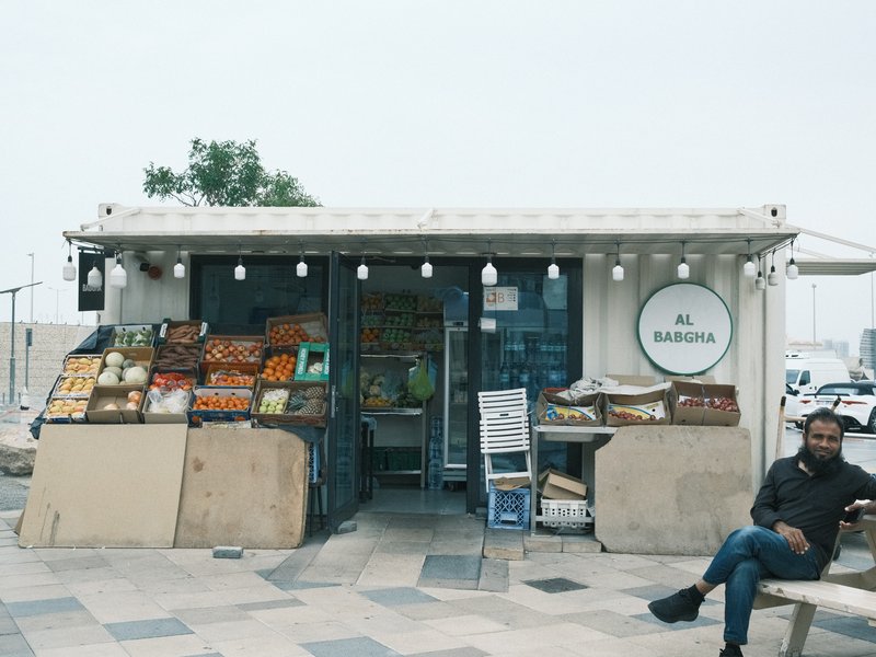 A small fruit and vegetable shop with a man sitting outside, surrounded by fresh produce.