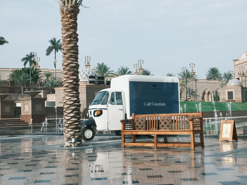 A food truck parked beside a palm tree on a wet pavement, with a bench in the foreground.