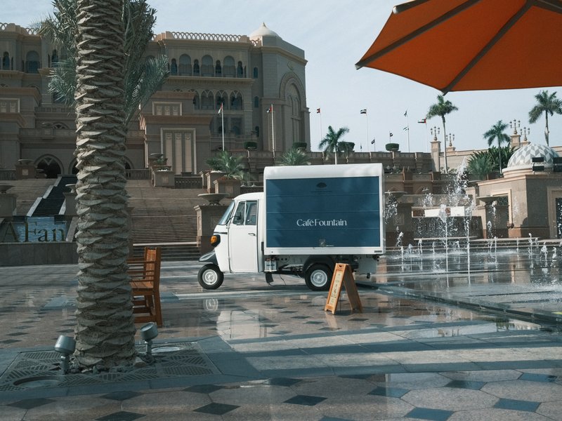 A small delivery truck parked near a fountain in a sunny outdoor plaza with palm trees and a large building.