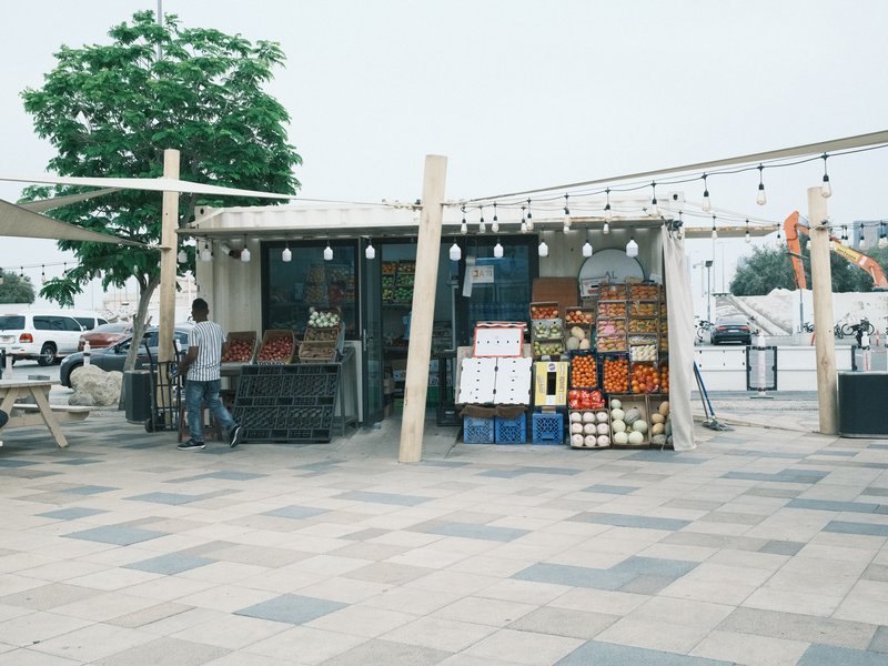 A small market stall with colorful produce and crates, set in an urban plaza with a tree and string lights.