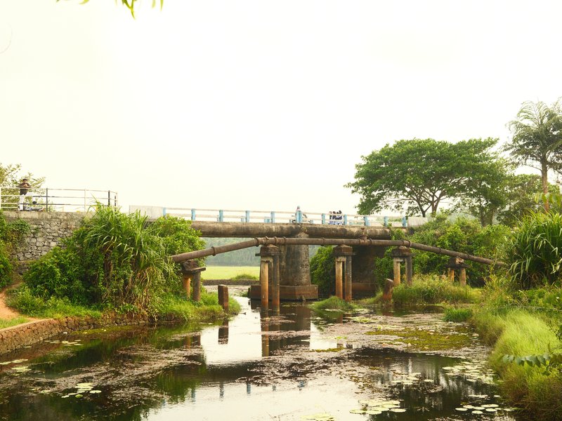 A rustic bridge over a calm waterway surrounded by lush greenery under a hazy sky.