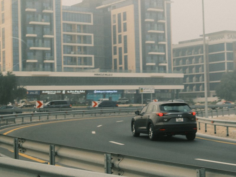 A black car driving on a curved road near modern buildings in a hazy urban setting.