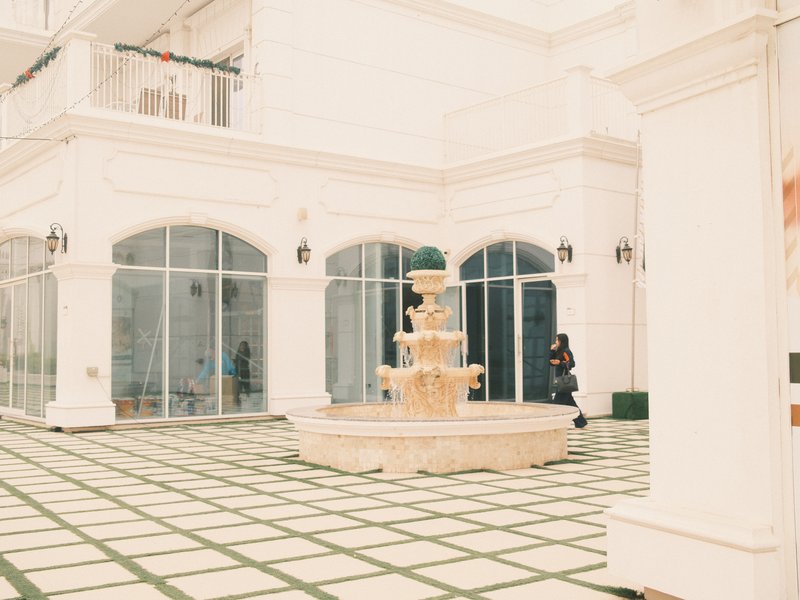 A serene courtyard featuring a decorative fountain surrounded by white architecture and large glass windows.