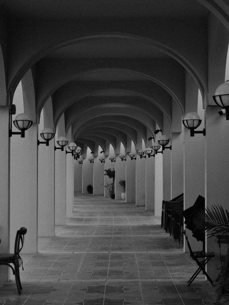 A monochrome corridor with arches and lamps, featuring benches along the sides.