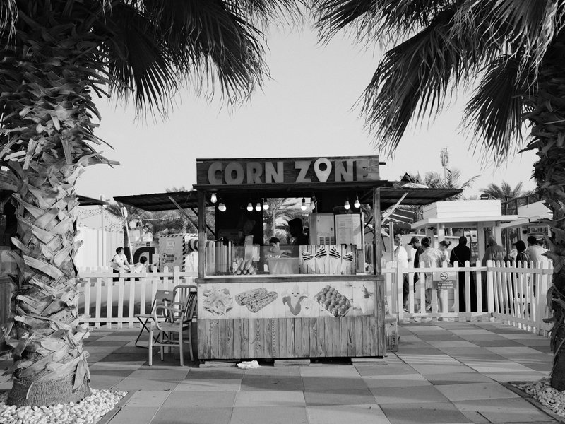A corn stand named 'CORN ZON' surrounded by palm trees and people in a lively outdoor setting.