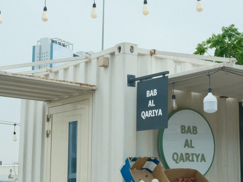 A modern container-style building with a sign reading 'Bab Al Qarya' and decorative lights overhead.