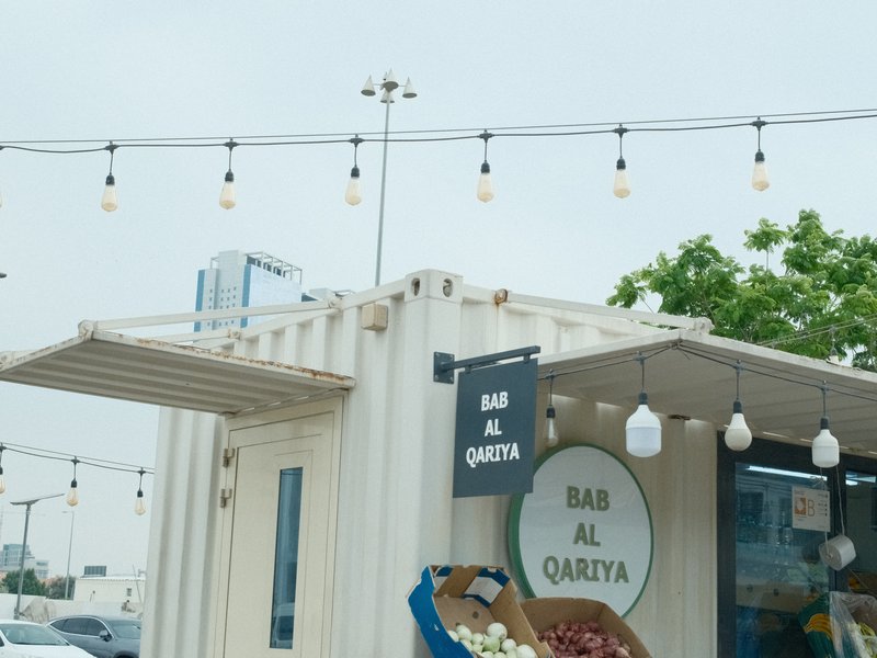 A small market stall with a sign reading 'Bai Al Qarya', surrounded by string lights and greenery.